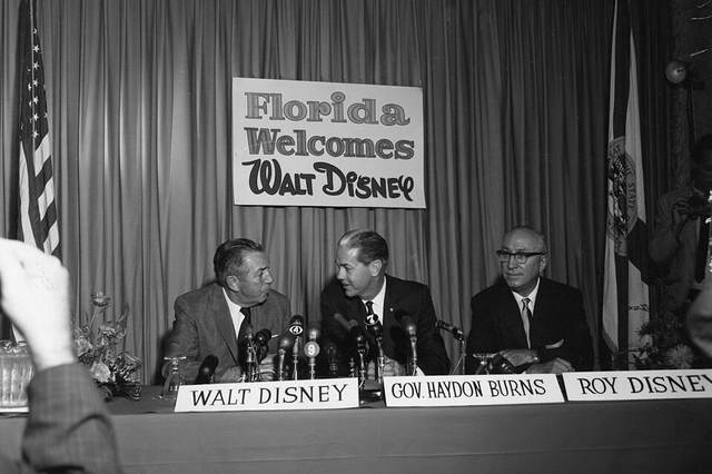 Walt Disney, Florida Gov. Haydon Burns and Roy O. Disney seated at a table of microphones under a sign reading Florida Welcomes Walt Disney